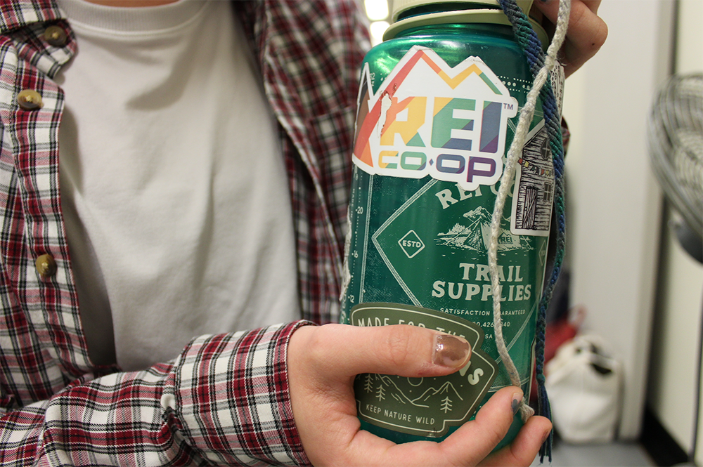 A person in a white t-shirt and flannel, head not pictured, holds a reusable, green plastic water bottle with a variety of stickers.
